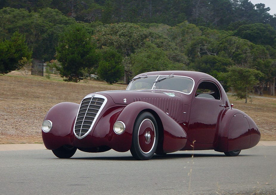 Alfa Romeo 8C 2300 coupé aerodinámico color vino, en carretera junto al bosque de Pebble Beach.