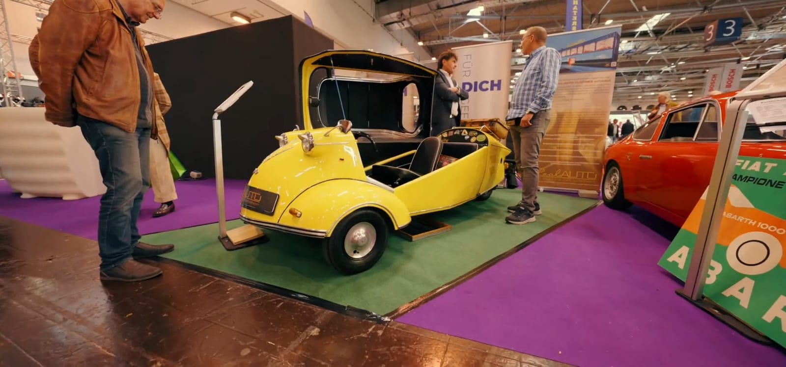 Microauto amarillo de tres ruedas con la cabina abierta, sobre alfombra verde.